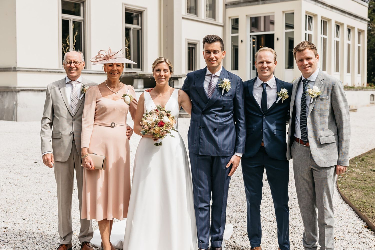 Wedding couple poses with four family members outside a grand estate on a sunny day.