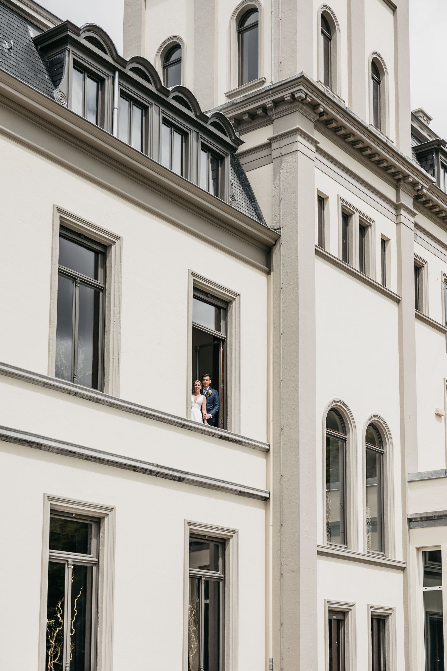 Bride and groom stand together in open window of elegant white neoclassical wedding venue building.