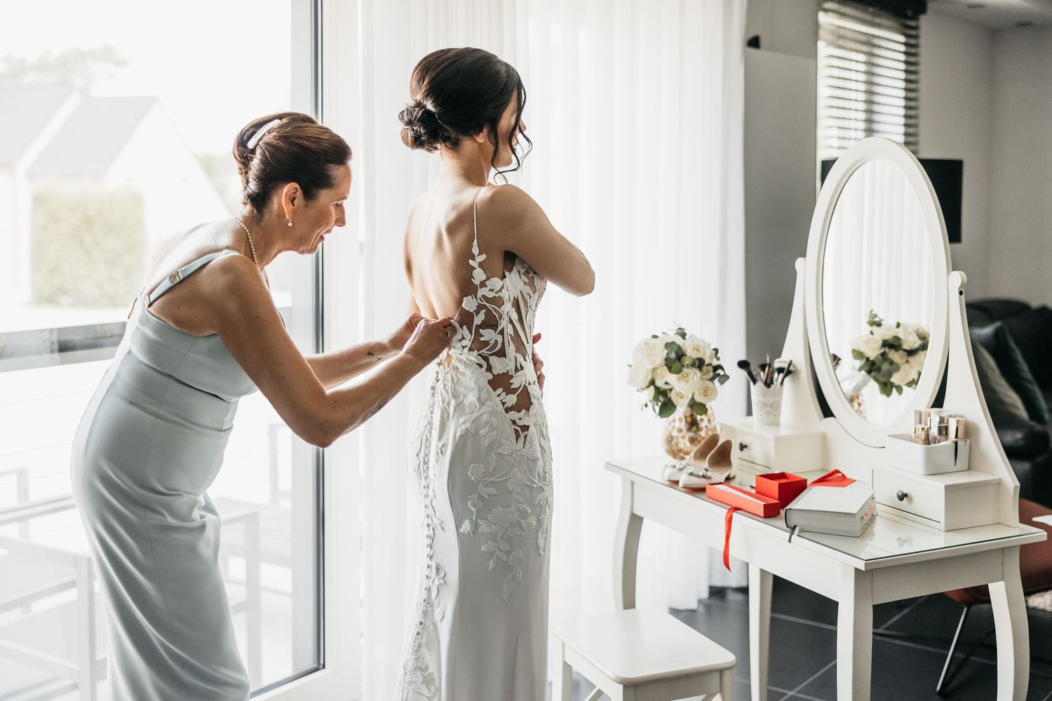 Woman assisting bride with lace wedding gown at a white vanity table.