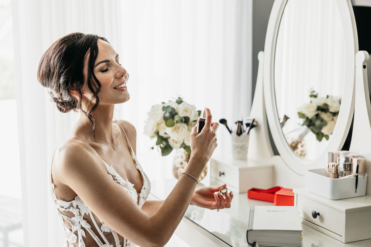 Radiant bride smiling while spraying perfume at a white dressing table on her wedding morning.
