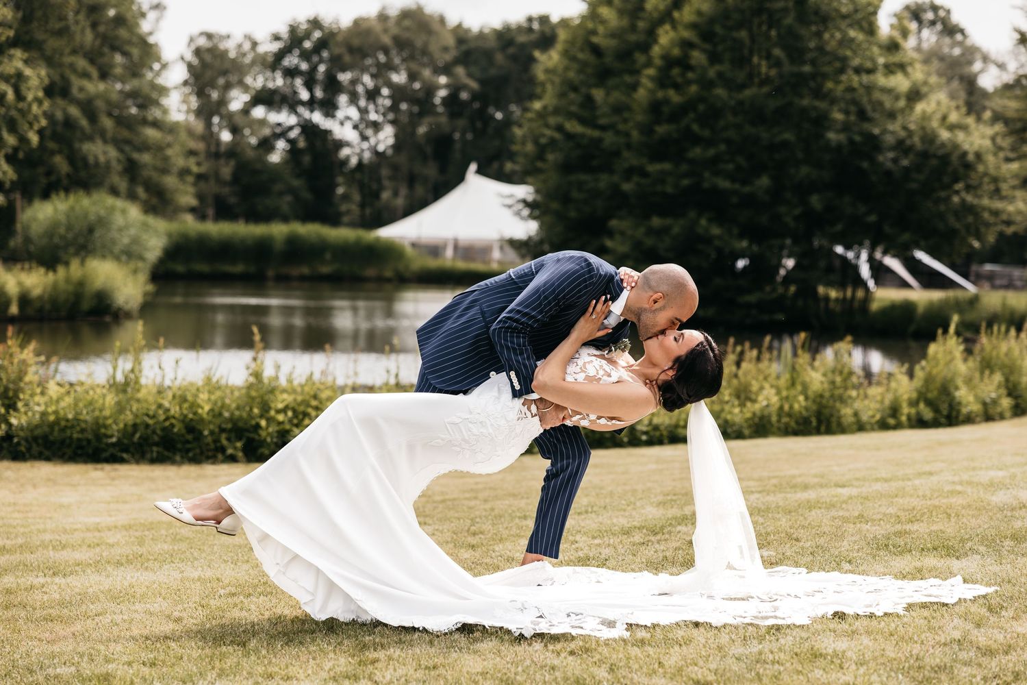 Groom dips bride in white gown for a romantic kiss on a sunny lawn with a marquee in the background.