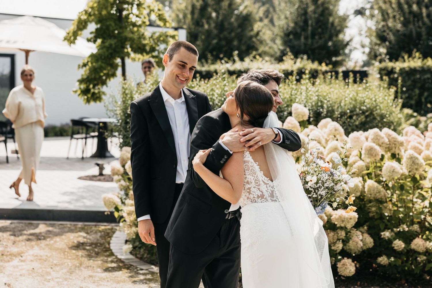 Groom smiles warmly while embracing wedding guest beside blooming white hydrangeas at outdoor ceremony.