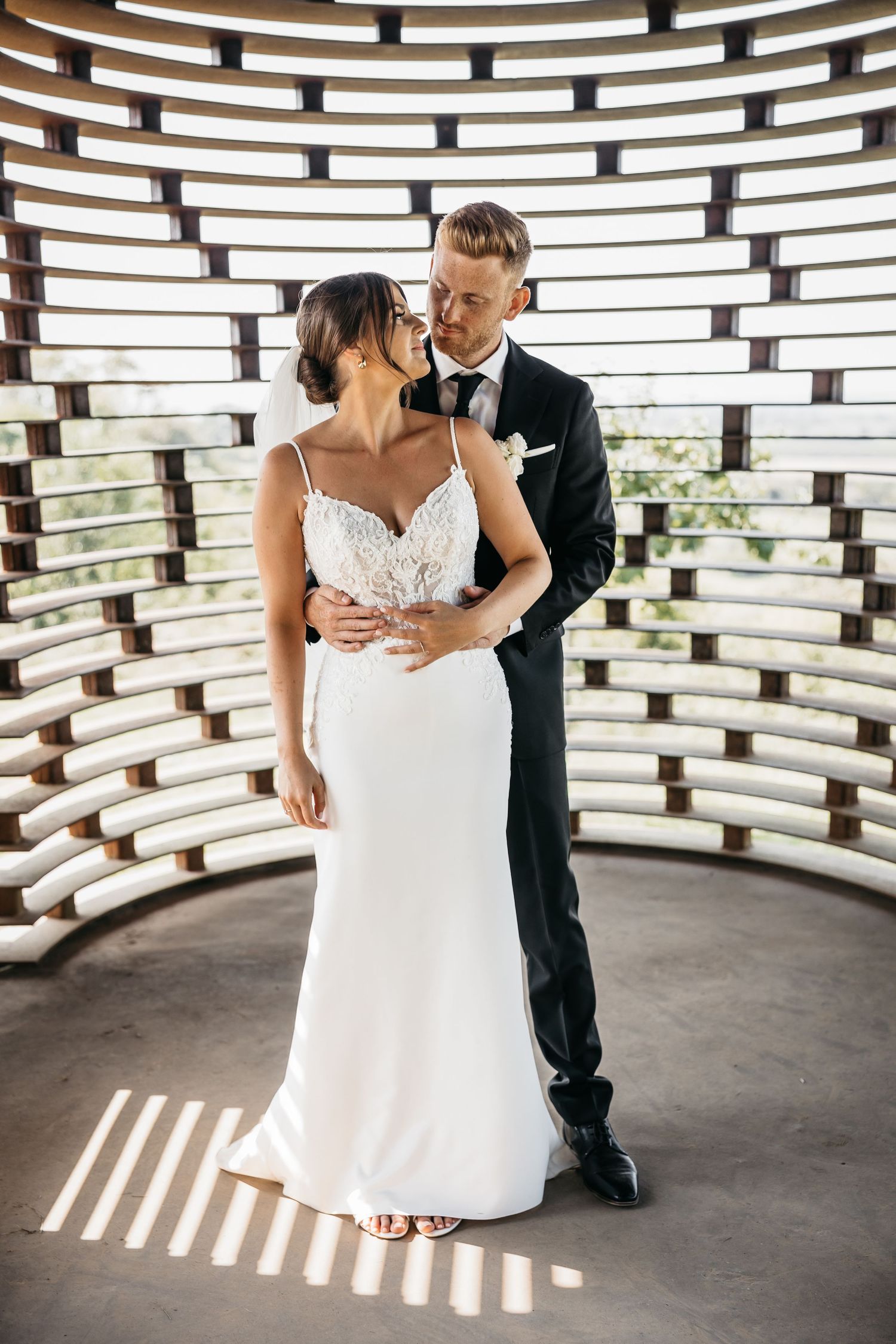 Groom embraces bride in a lace wedding dress inside a stunning curved wooden lattice chapel on their wedding day.