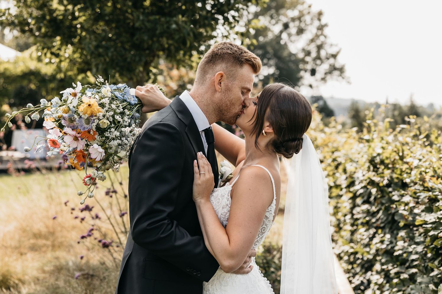 Newlyweds sharing a tender kiss outdoors surrounded by lush greenery and a vibrant wildflower bouquet.