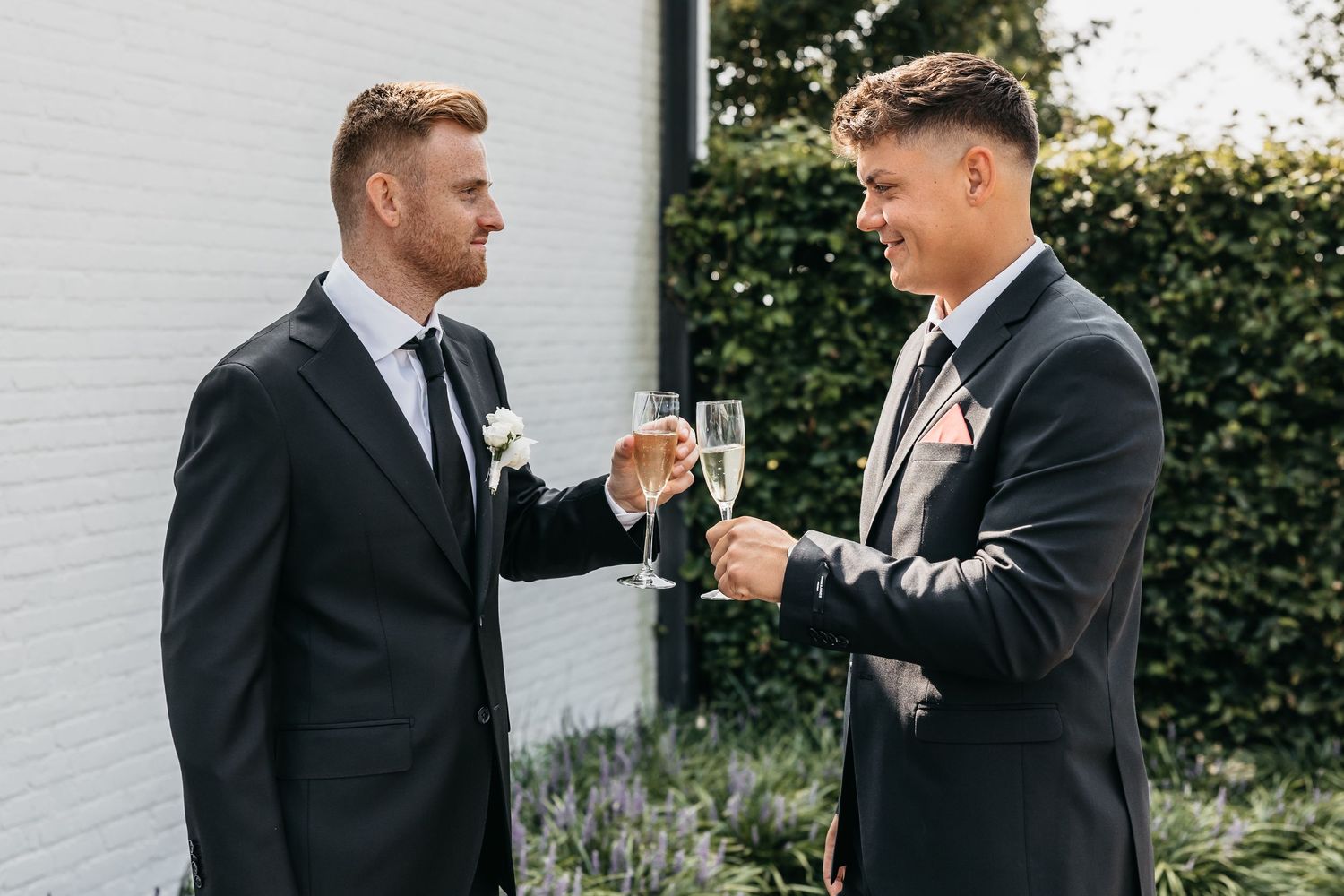 Two groomsmen in black suits clink champagne glasses and smile outdoors at a wedding.