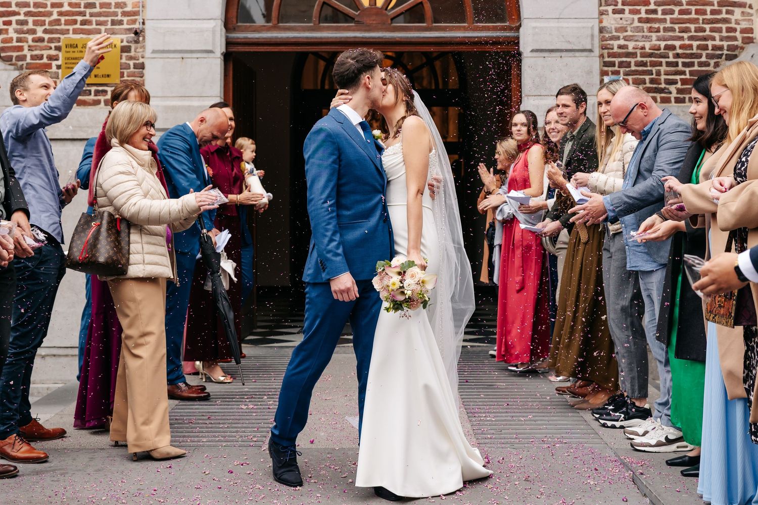 Bride and groom share a kiss outside church while guests line up to celebrate with confetti.