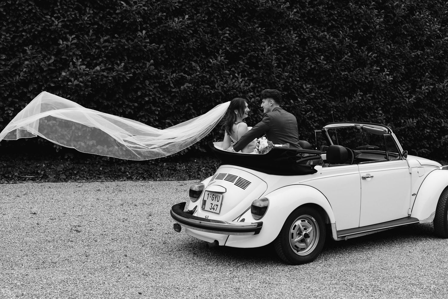 Black and white photo of newlyweds kissing in vintage VW Beetle convertible with veil blowing in wind.