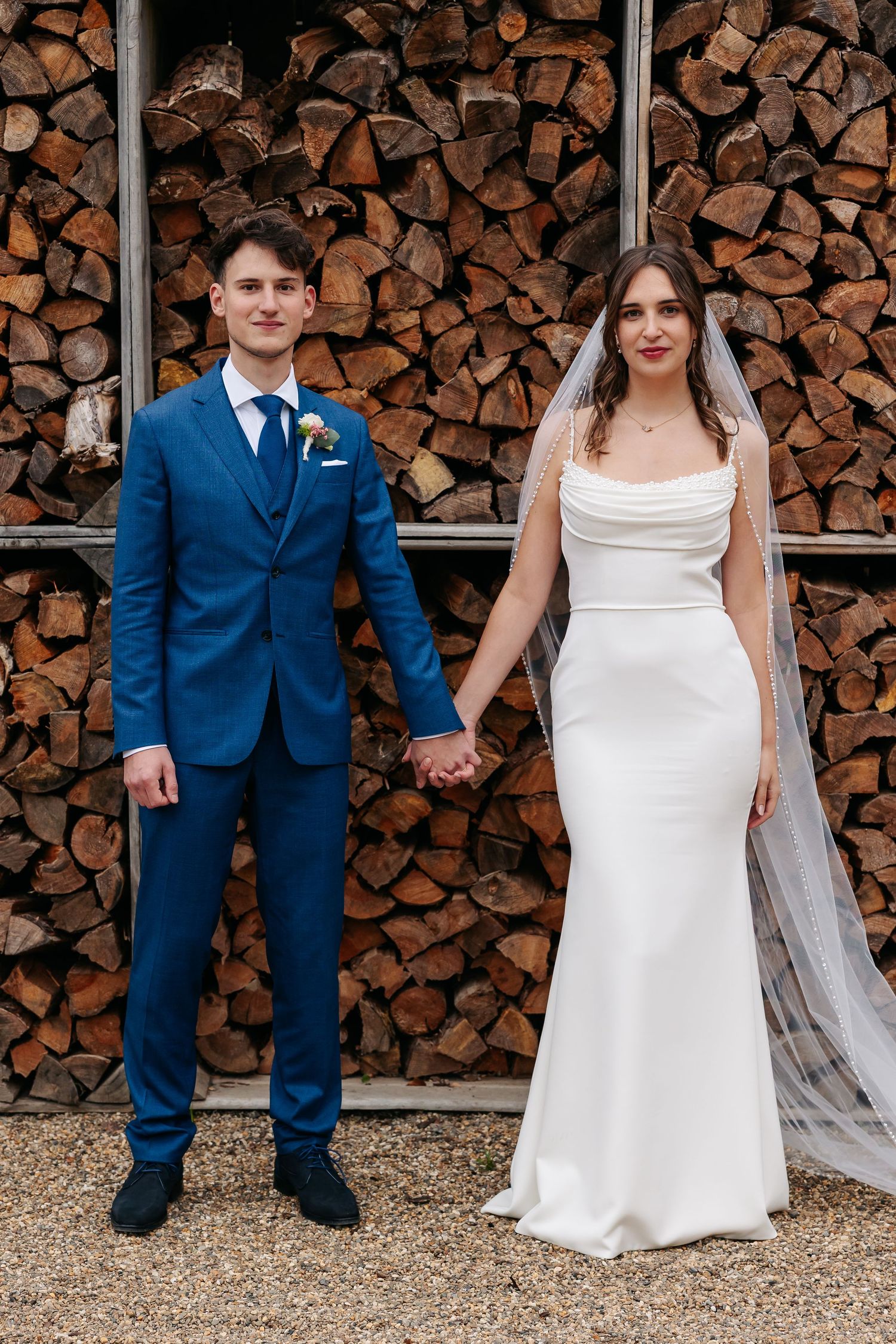 Bride in white gown and groom in blue suit holding hands before a rustic stacked firewood backdrop.