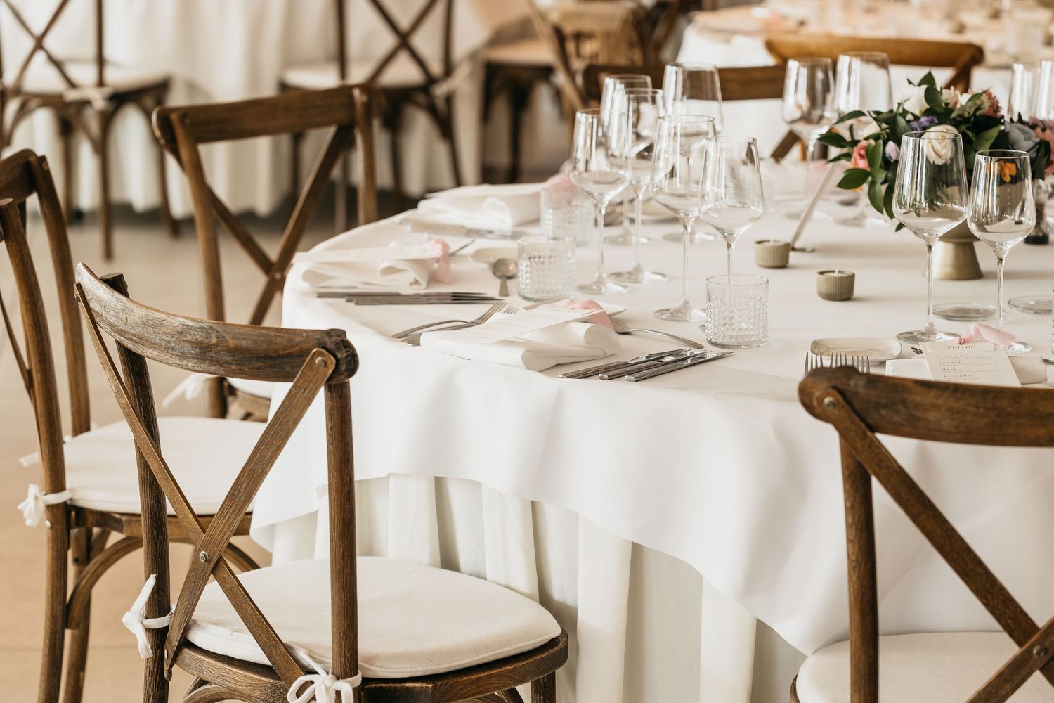 Wedding reception table with white linen, wooden cross-back chairs, glassware, and floral centerpiece.