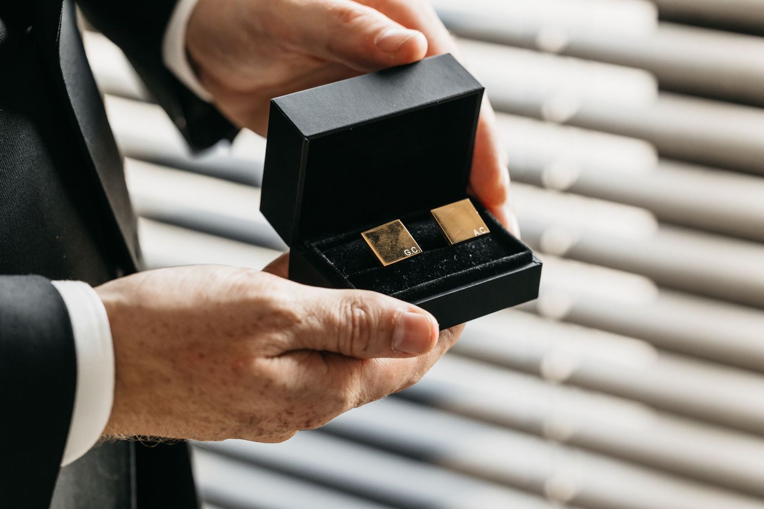 Groom holds open black velvet box displaying gold square cufflinks as a wedding day gift.