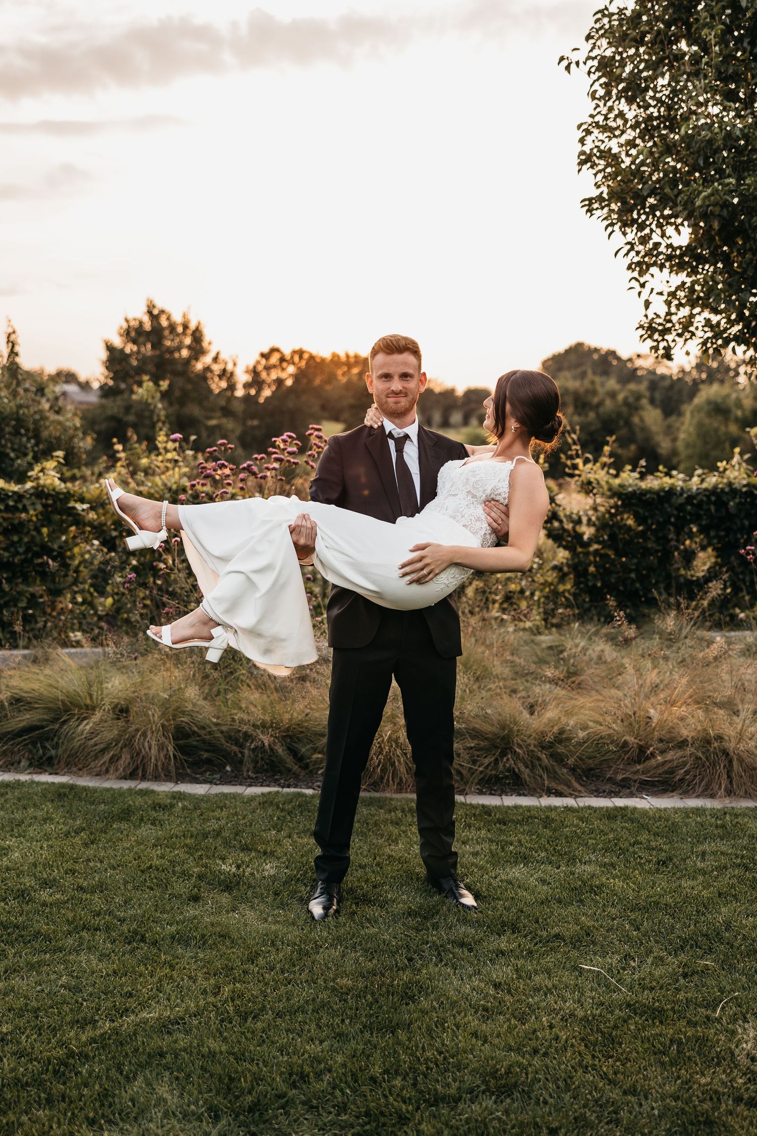 Smiling groom in black suit carries bride in white wedding dress in his arms against a lush garden sunset backdrop.