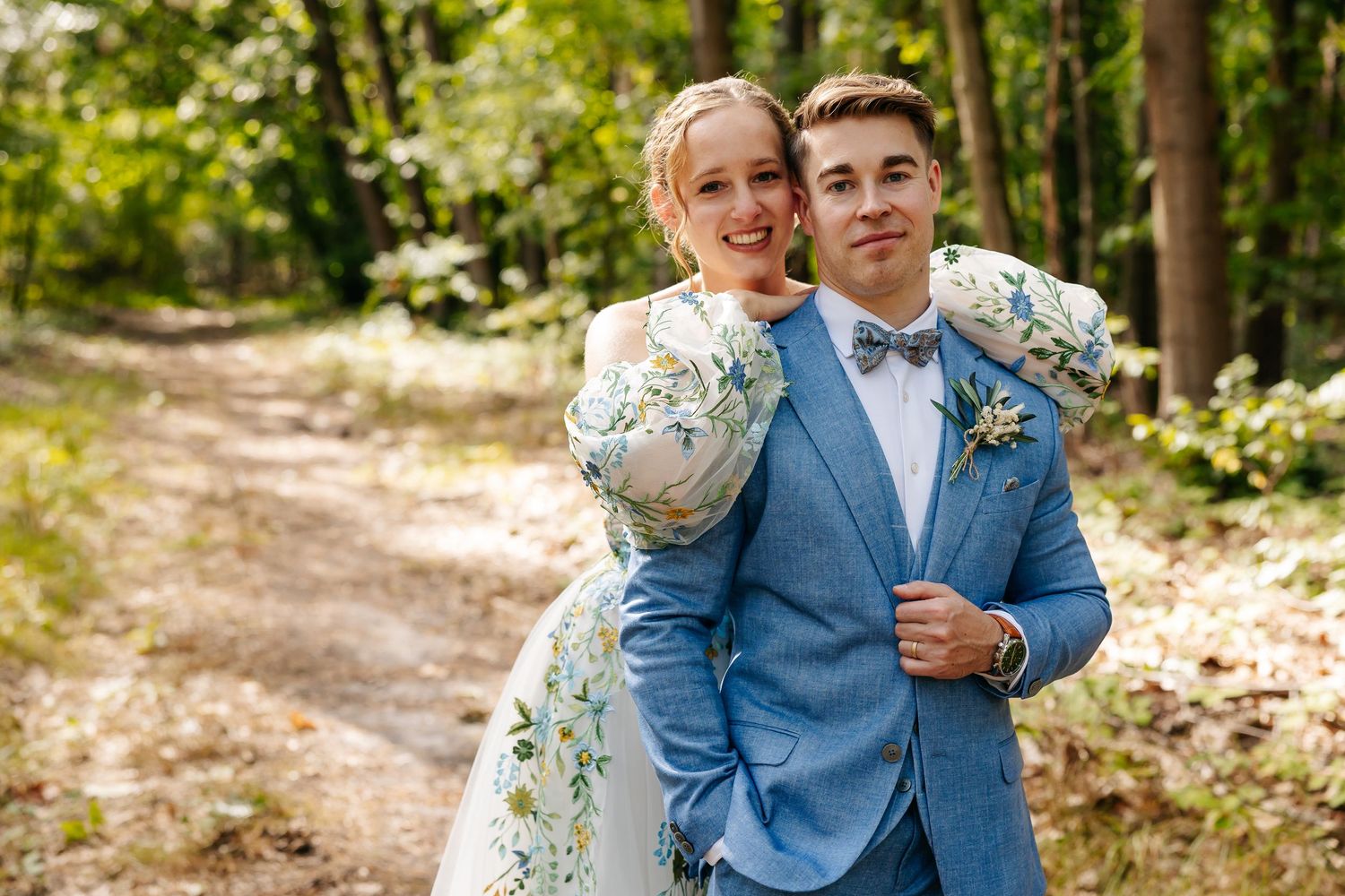 Happy bride in floral gown embracing groom in blue suit with bow tie on a forest path.
