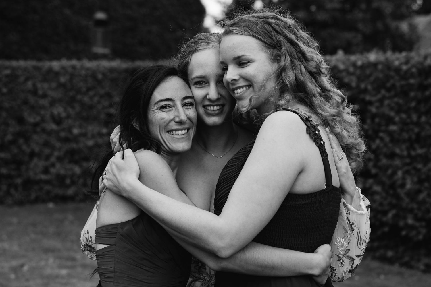 Black and white photo of bride hugging two bridesmaids in joyful embrace in garden setting.