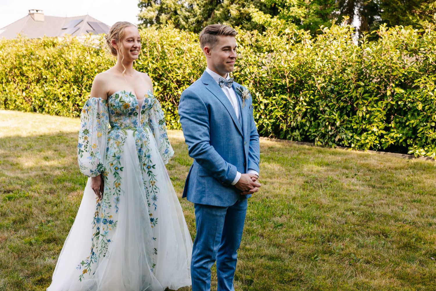 Bride in elegant floral wedding gown stands beside smiling groom in light blue suit outdoors.