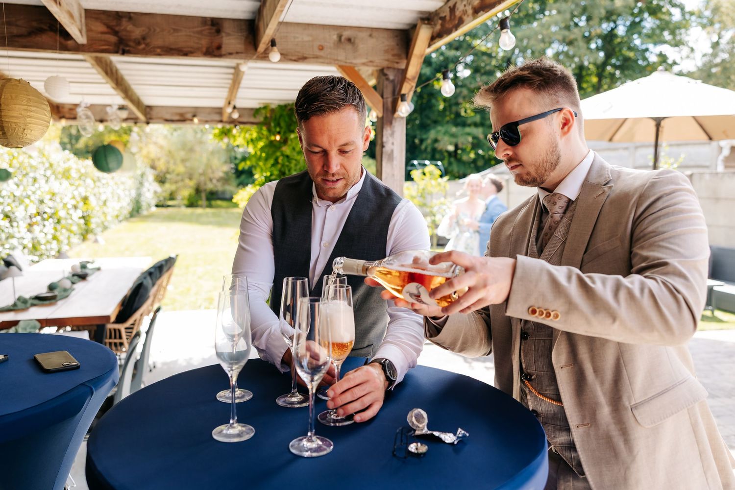 Two wedding guests pour champagne into glasses at elegant blue-draped cocktail table.
