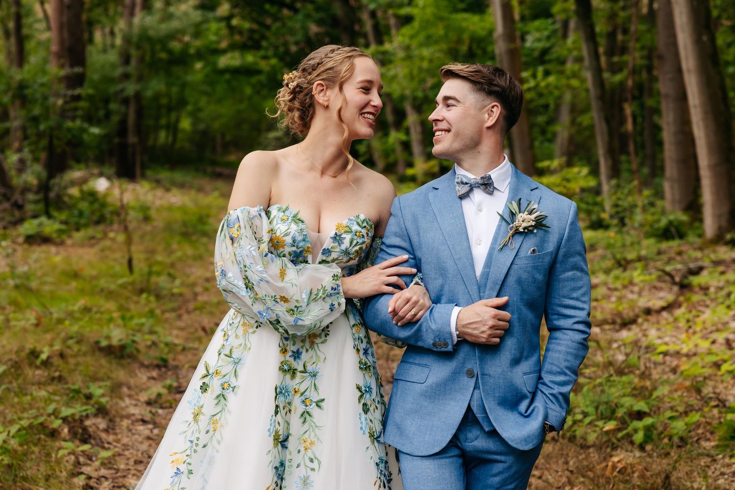 Smiling bride and groom in blue suit embracing in green forest.