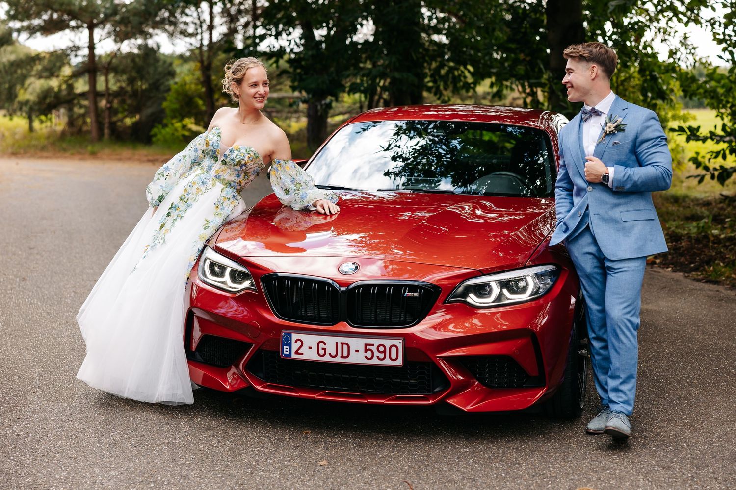 Bride and groom posing with red BMW M2 on their wedding day outdoors.
