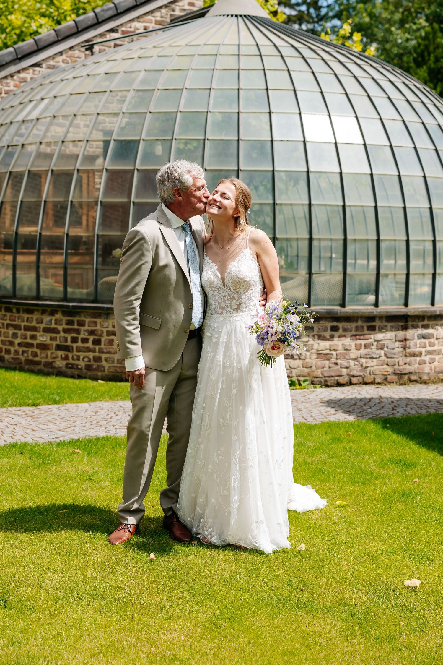 Bride and groom sharing a kiss in front of a glass dome greenhouse on a sunny wedding day.