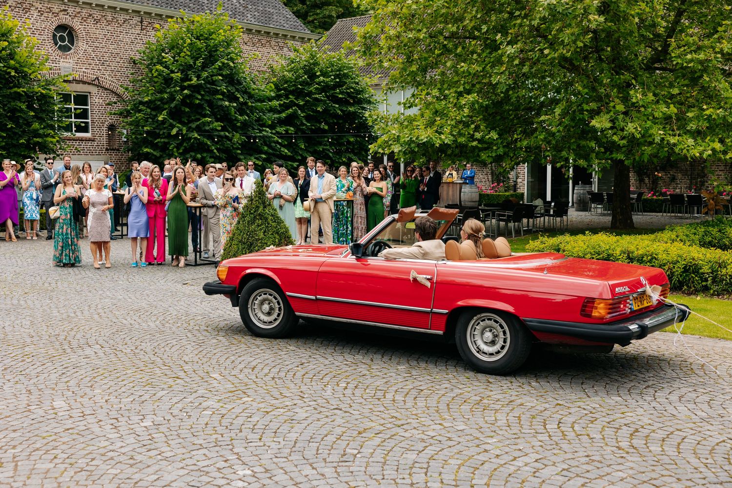 Newlyweds driving away in a classic red Mercedes convertible past cheering wedding guests.