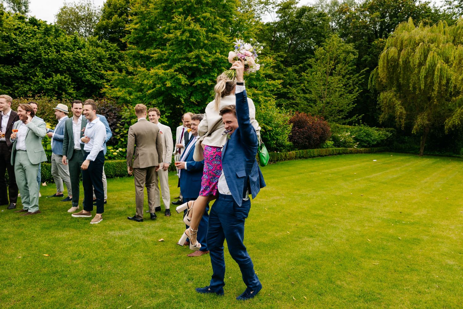 Wedding guest playfully lifts bride while holding bouquet high on green lawn at outdoor celebration.