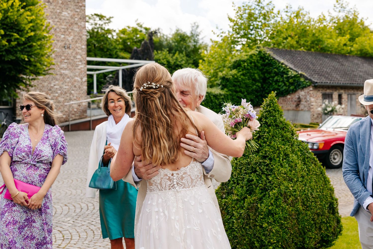 Bride in white lace wedding dress embracing a guest outdoors at a rustic countryside wedding venue with flowers.