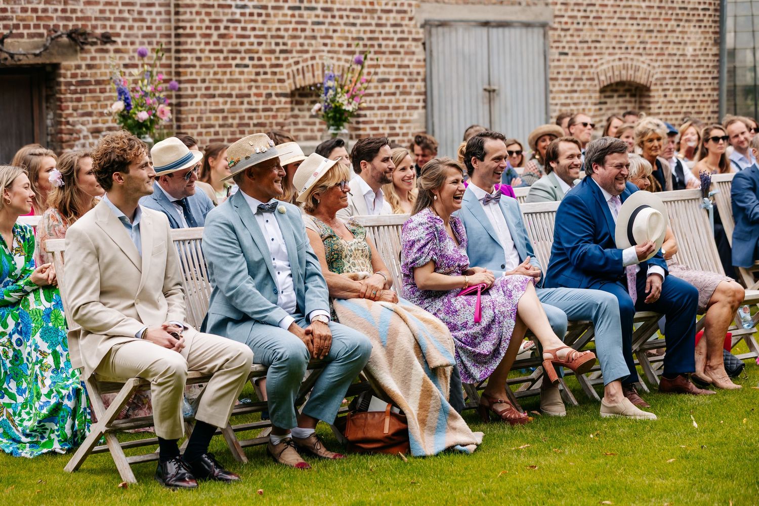 Wedding guests seated outdoors in summer attire watching ceremony at rustic brick estate venue.