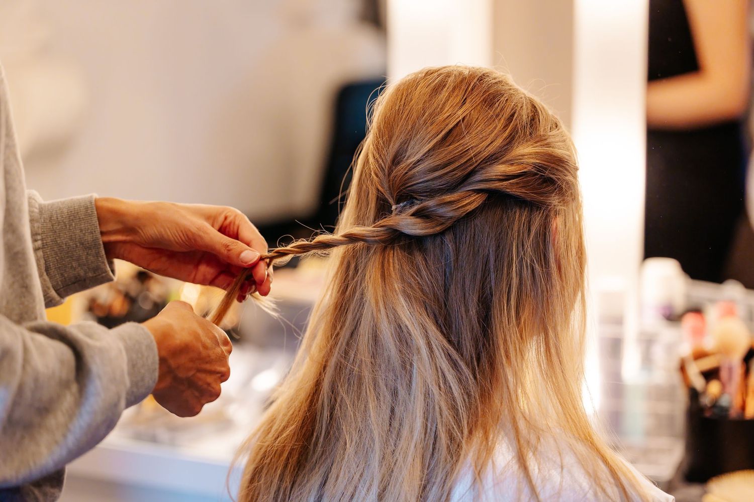 A hairstylist braiding a blonde woman's hair in a salon mirror setting.
