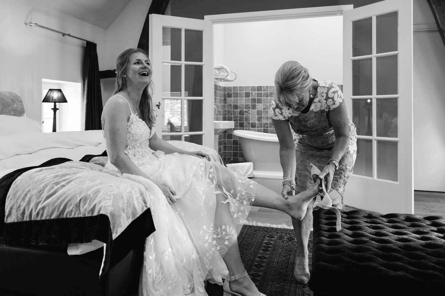 Bride laughing as bridesmaid helps put on wedding shoes in elegant black and white photo.