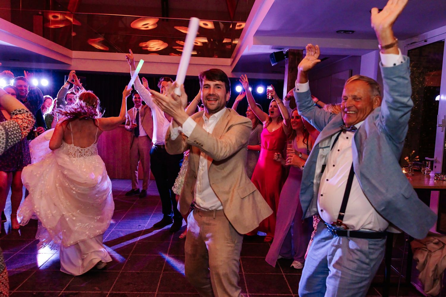 Groom in beige suit dancing with bride and older guest holding foam sticks at lively wedding reception.