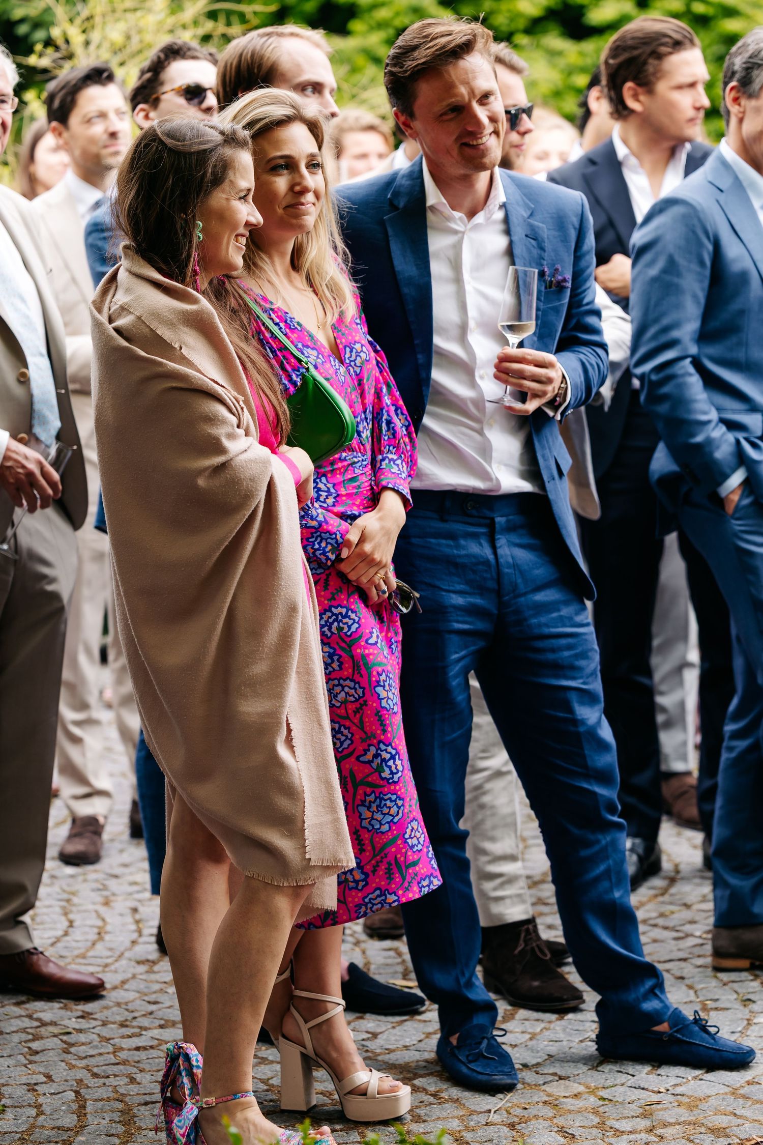 Wedding guests socializing outdoors, woman in pink floral dress standing with man in blue suit.