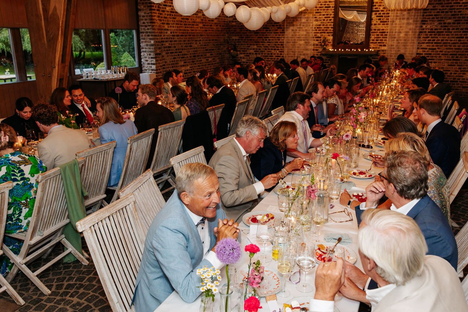 Large wedding reception dinner with guests seated at long tables in a warmly lit rustic brick venue.