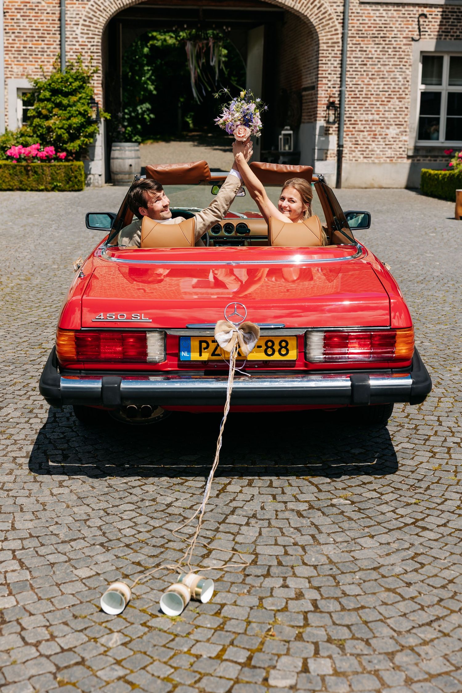 Newlyweds celebrating in a classic red Mercedes convertible with tin cans trailing on cobblestone driveway.