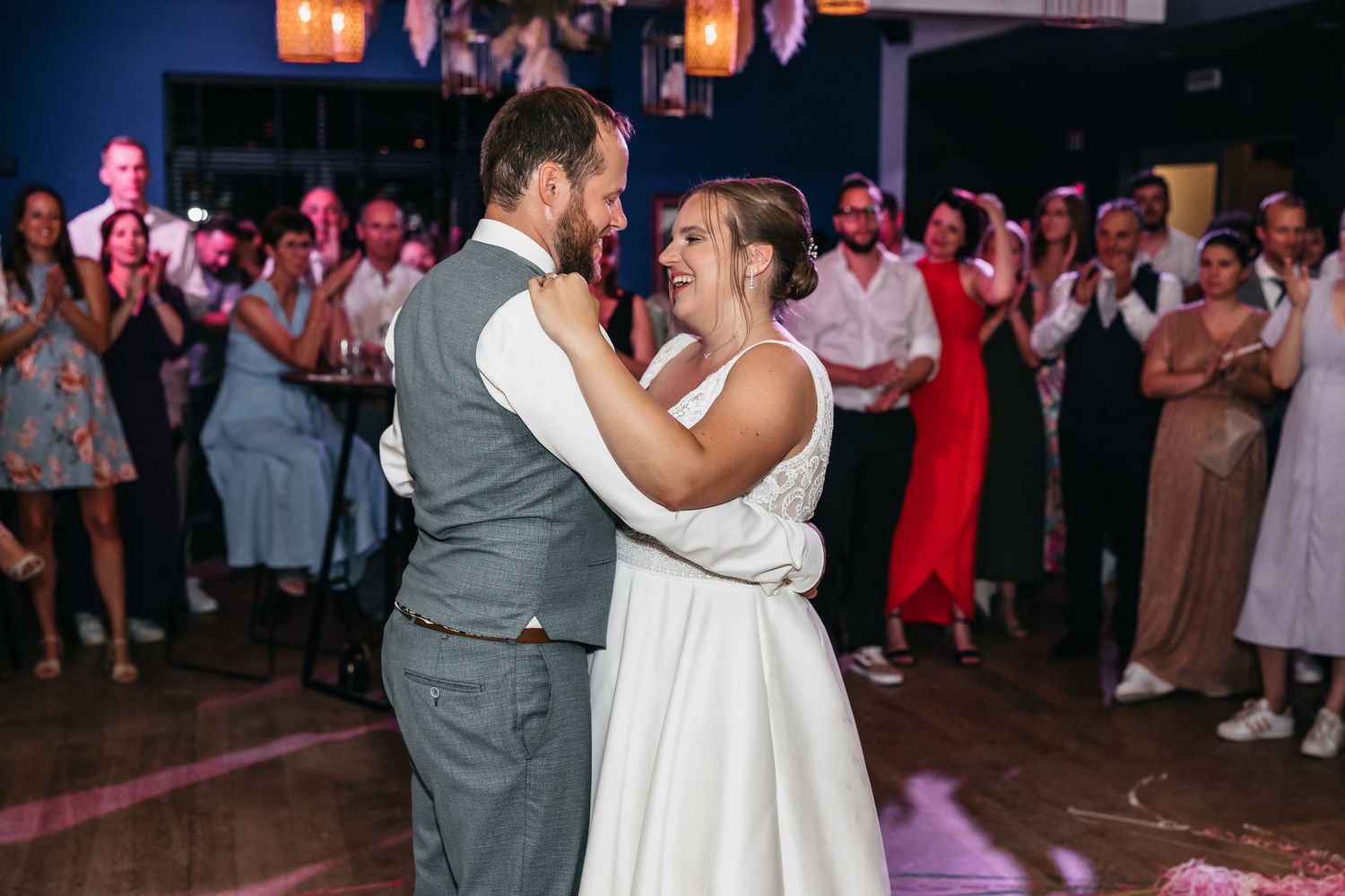 Happy bride smiles at her groom during their first dance as guests applaud at a vibrant wedding reception.