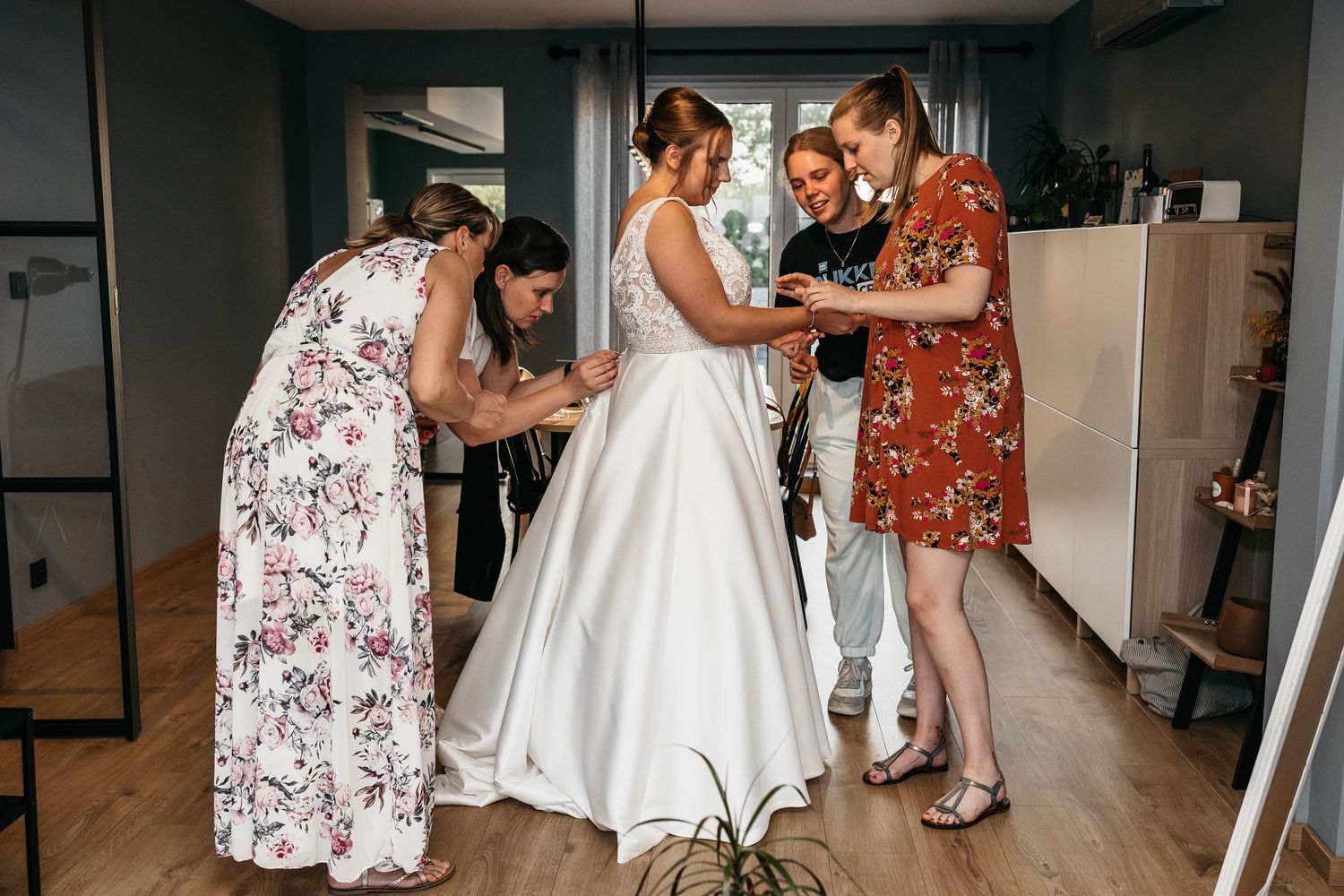 Bride in white satin gown being assisted by three women with dress details in modern interior.