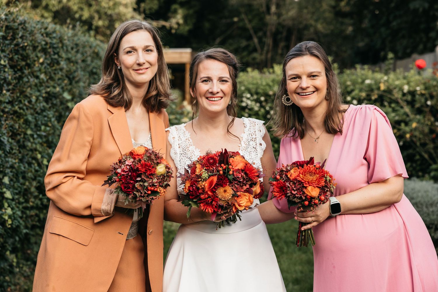 Bride and two bridesmaids smiling outdoors holding vibrant red and orange autumn wedding bouquets.