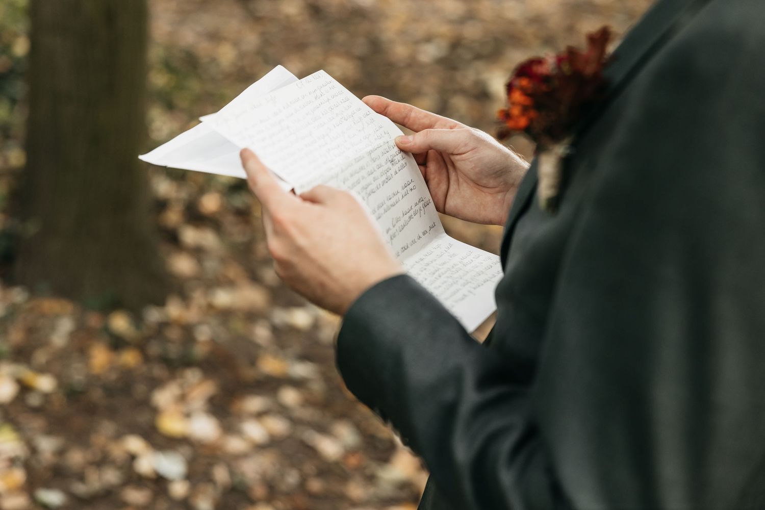 Groom in gray suit reading wedding vows on paper in an autumn forest setting.