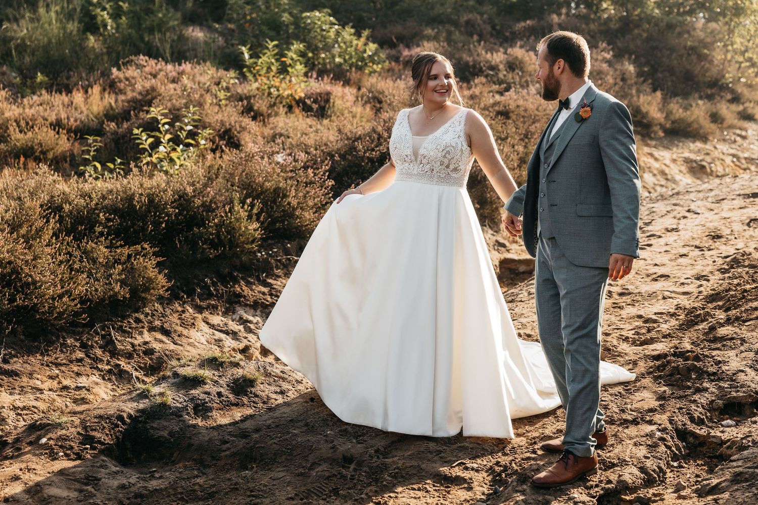Bride and groom walking hand in hand along a sandy path through a golden heather landscape.