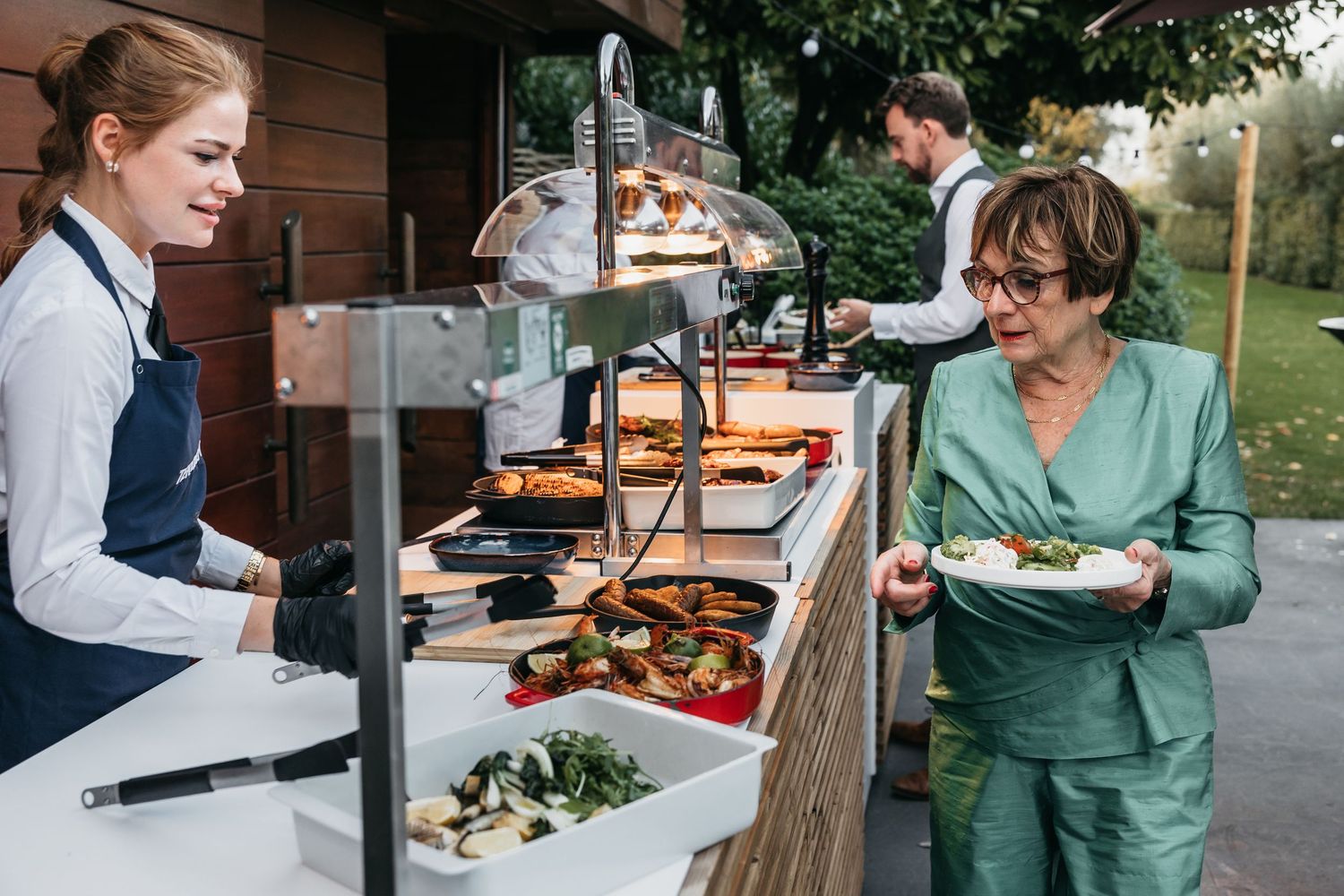 Female caterer serving grilled vegetables to a guest in green suit at an outdoor buffet.