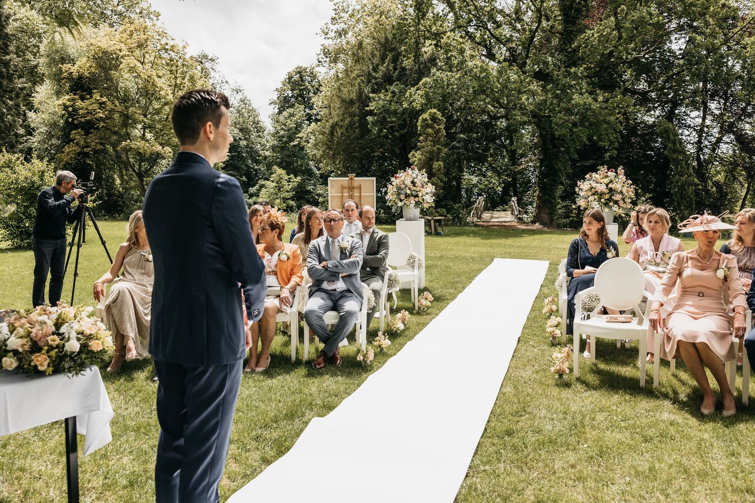 Groom in navy suit waits at altar of elegant outdoor garden wedding ceremony with seated guests.