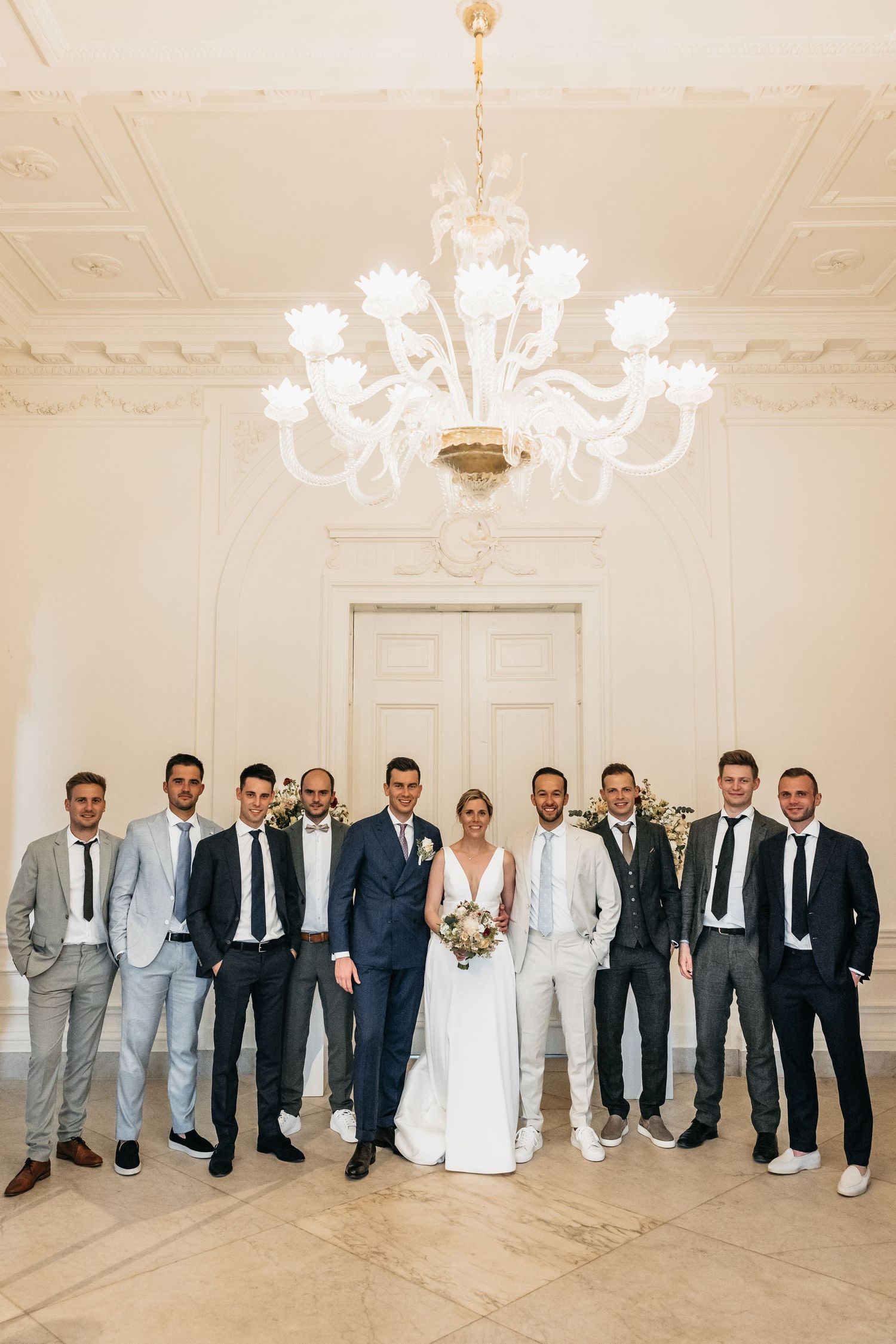 Bride and groom pose with groomsmen in suits under elegant chandelier in ornate white venue.