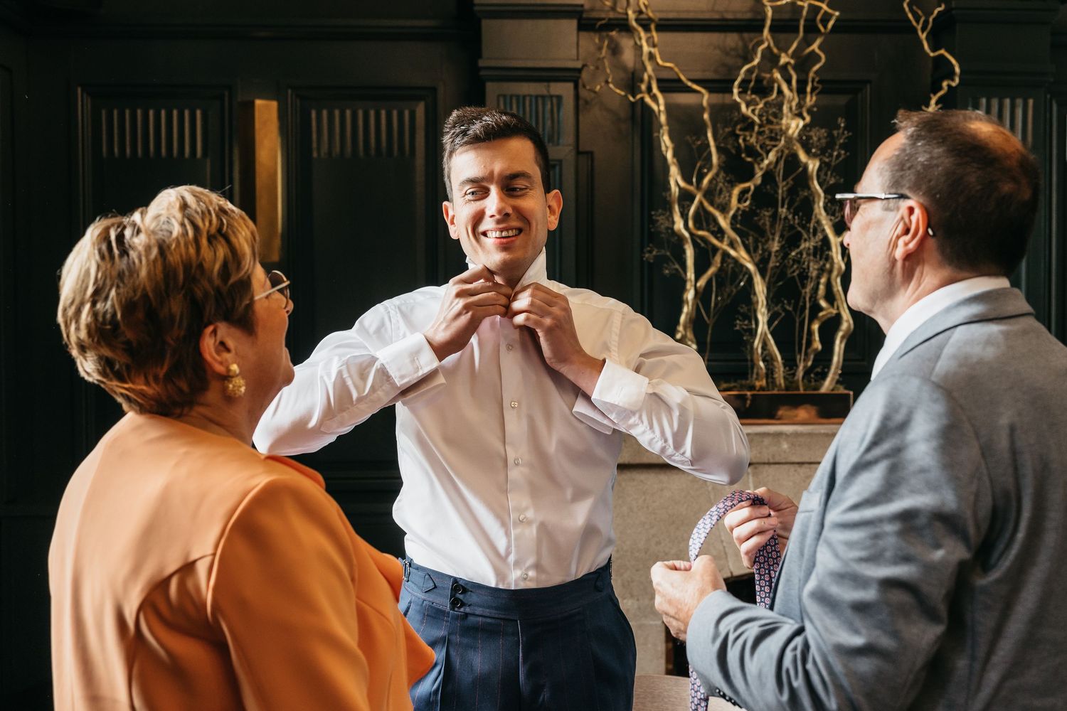 Groom in white shirt adjusting his collar while two guests look on in an elegant room.