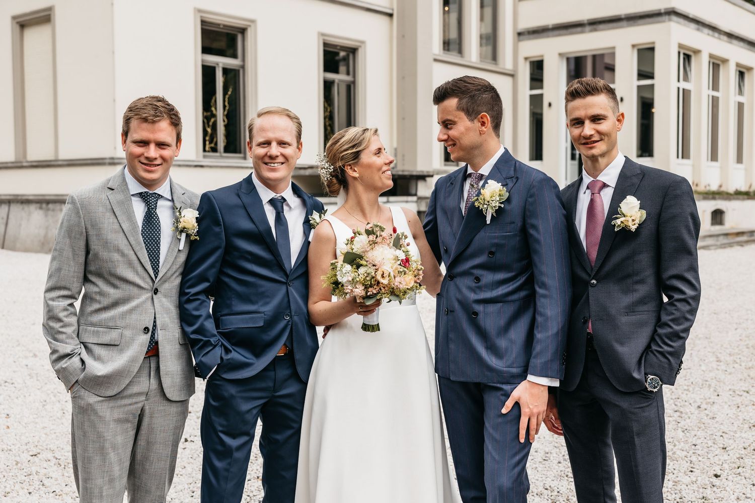 Wedding party of bride and groomsmen posing in navy suits outside elegant venue.
