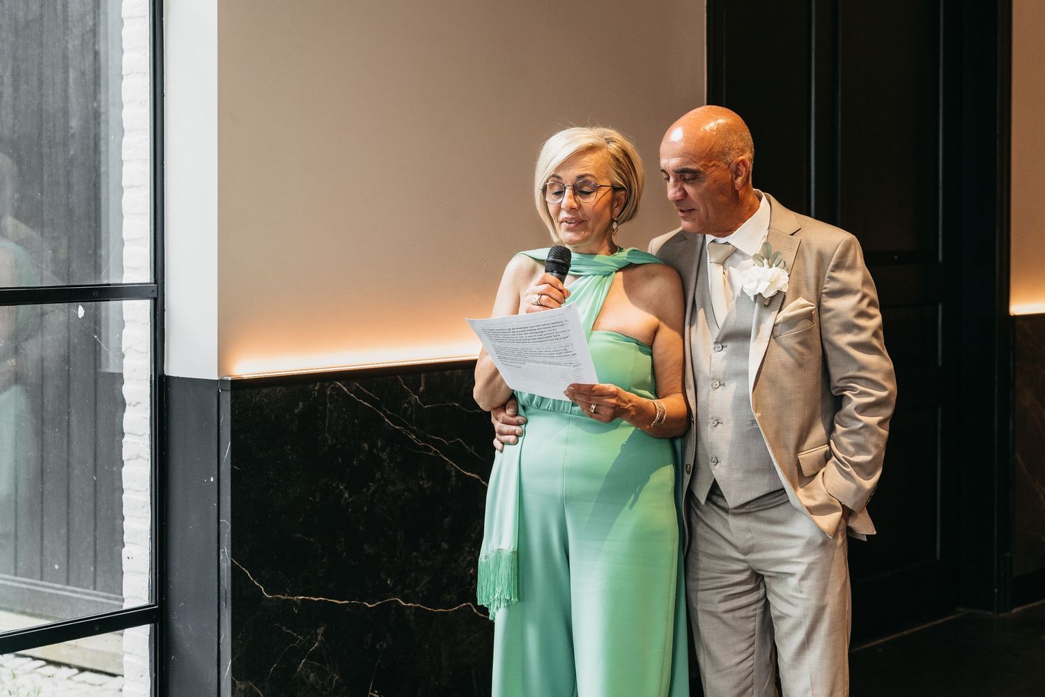 Older couple in formal attire delivering speech at wedding reception, woman holding microphone and paper.