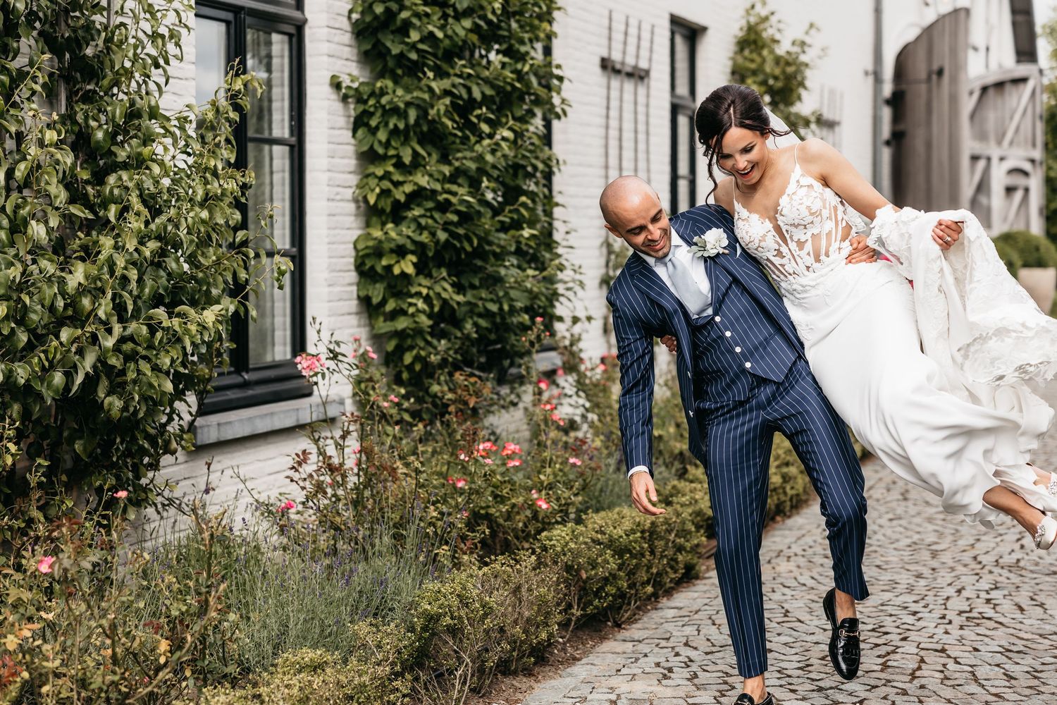 Groom in navy pinstripe suit playfully carries laughing bride in lace wedding dress on cobblestone path.