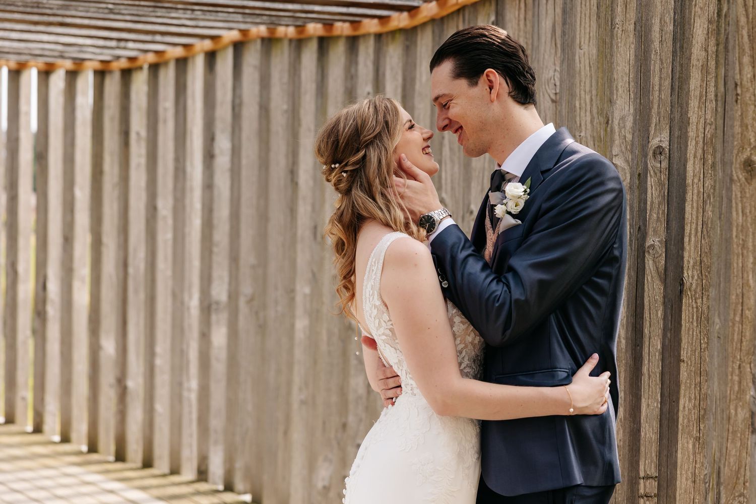 Bride and groom share a tender almost-kiss moment against rustic wooden wall outdoors.