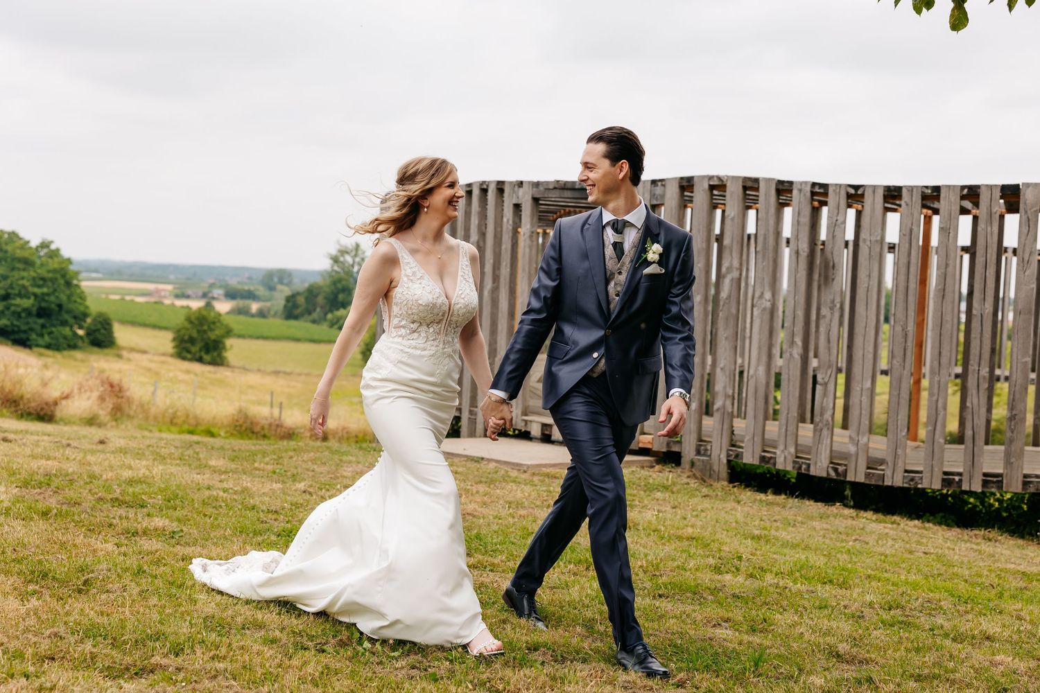 Smiling bride and groom holding hands while walking across a green lawn with a wooden fence in the background.