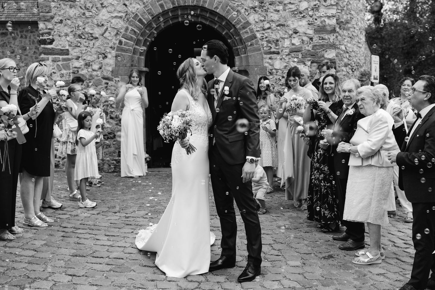 Black and white photo of newlyweds kissing outside stone church amid confetti-throwing guests.
