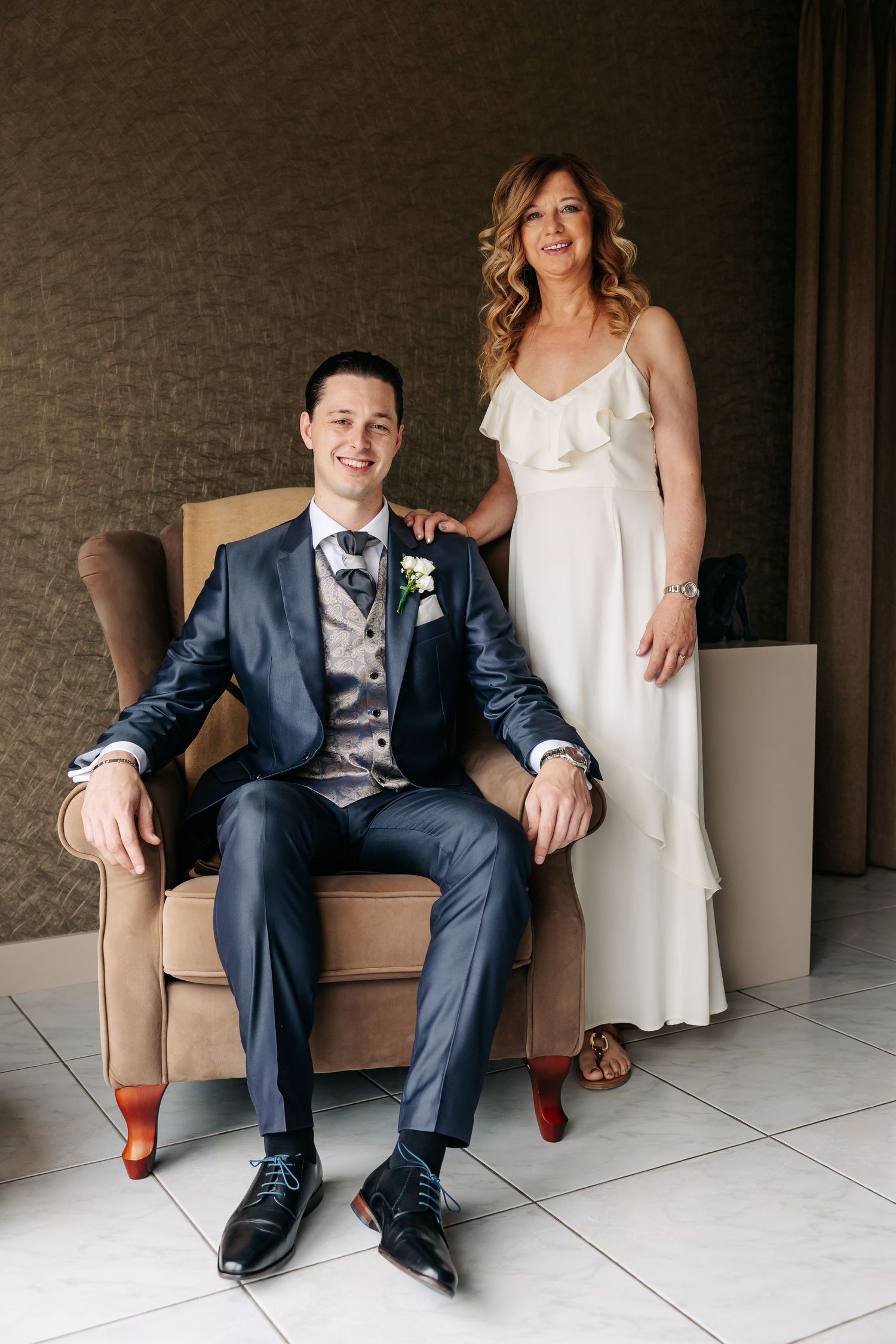 Groom in navy suit sitting in armchair posing with smiling woman in white dress.