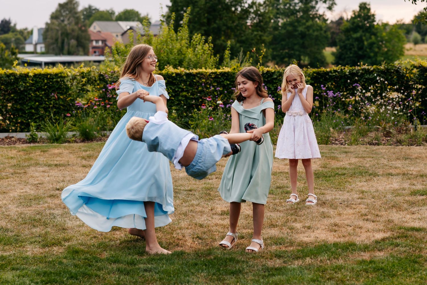 Bridesmaids in blue and green dresses laugh while swinging a child in a sunny garden at a wedding.