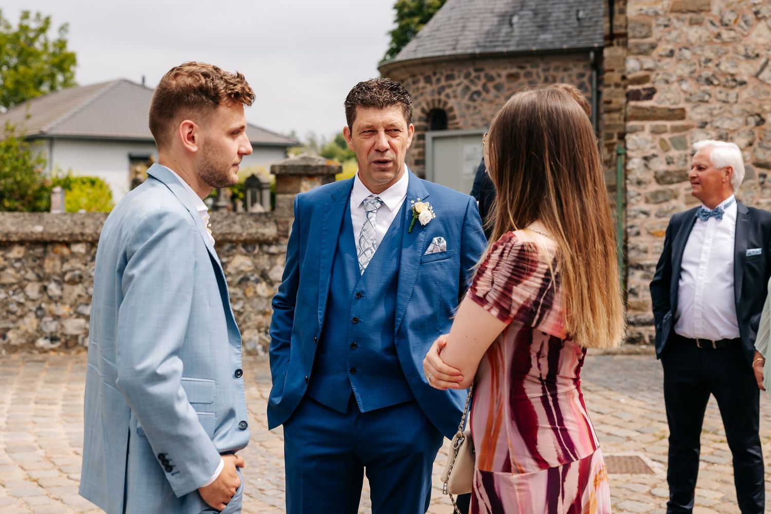 Wedding guests in formal attire conversing outdoors near stone building at a wedding reception.